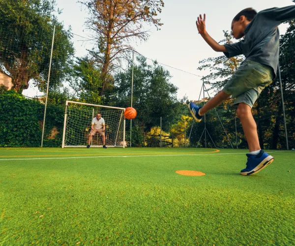 Young boy kicking a soccer ball towards a goal on a sunny day.