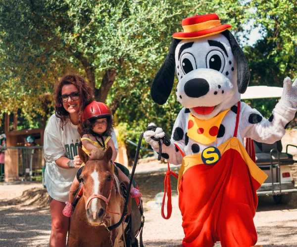 Child rides a pony with a woman; Spotty waves nearby.