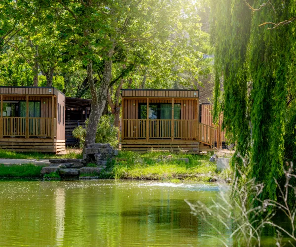Wooden cabins by a sunlit, tree-lined pond.
