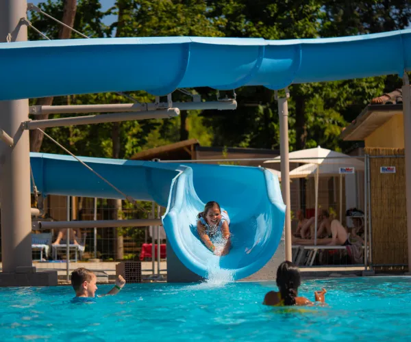 Child slides down blue water slide into a pool with other children swimming.