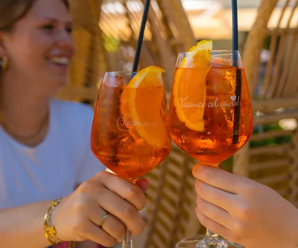 Two people clinking glasses with orange drinks, one smiling woman in the background.