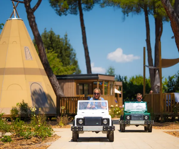 Two people driving mini cars past glamping tents and trees.