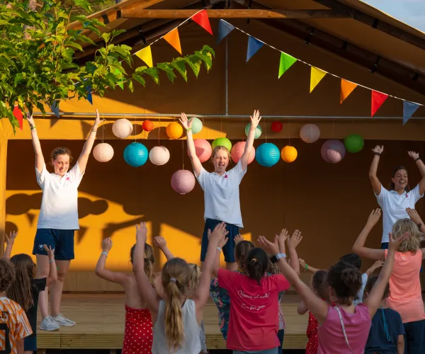 Children and instructors dance energetically under colorful bunting on a stage.