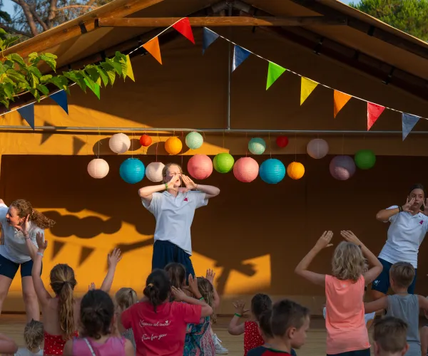 Outdoor stage with performers dancing, colorful flags, and a crowd of children watching.