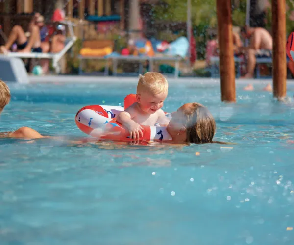 Toddler with floaties plays in a pool, surrounded by adults and kids.