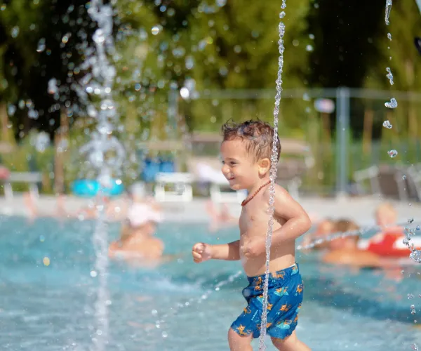 Young boy running through water jets at a poolside splash pad.