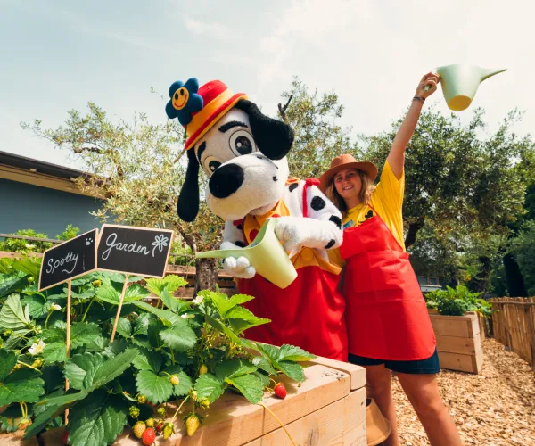 Dog mascot and person watering a garden, both smiling.