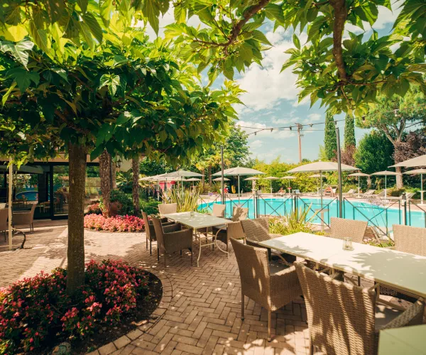 Sunny patio with tables near pool, surrounded by lush greenery and flowers.