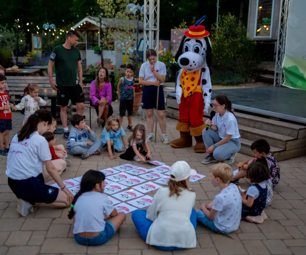 Children and a costumed character play a game outdoors, sitting on the ground in a circle.