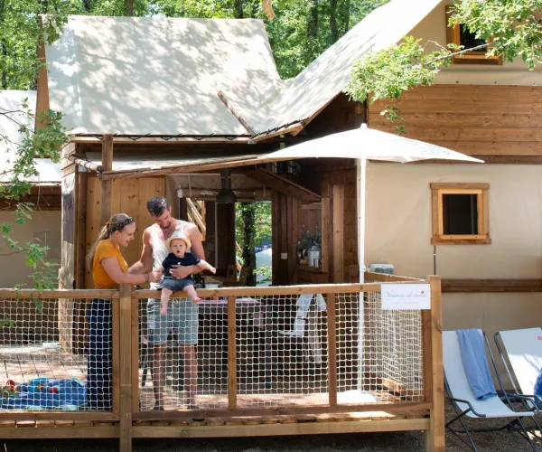 Family on a cabin porch surrounded by trees, with lounge chairs nearby.