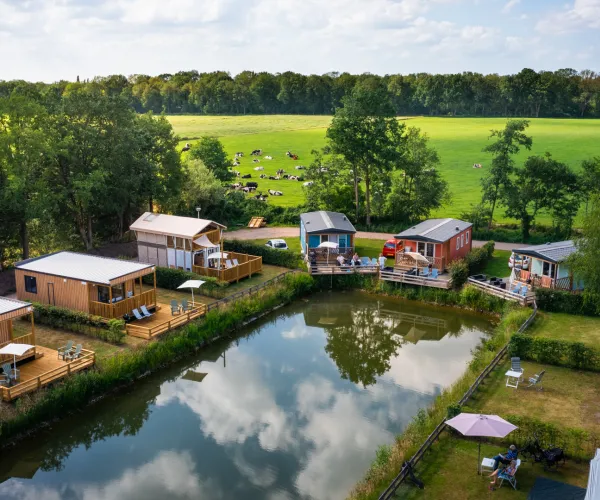 Cabins by a pond, surrounded by grass and trees, under a partly cloudy sky.