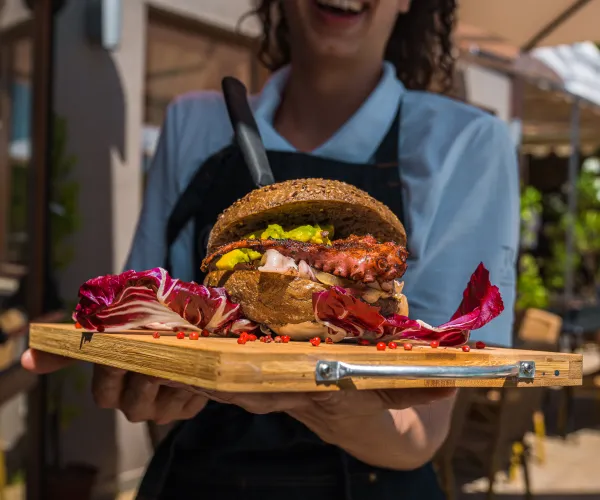 Smiling server holds a gourmet burger on a wooden platter outdoors.