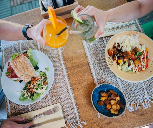 Two people clinking drinks over plates of salad and sandwiches.