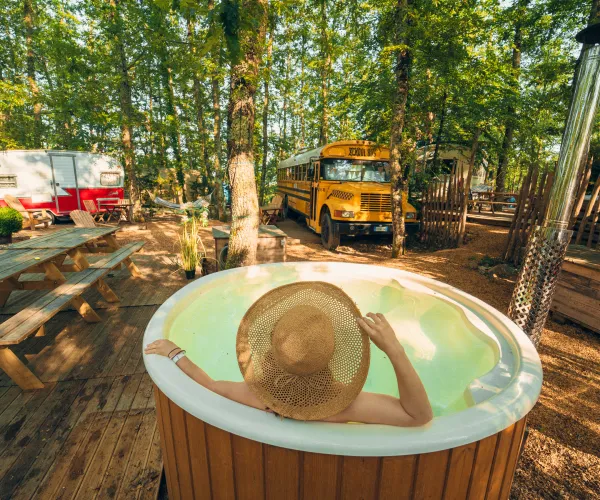 Person in a hat relaxing in a forest hot tub, with a bus and camper nearby.
