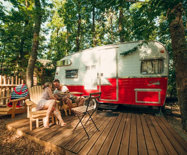 Camper in a wooded area with people sitting on a deck.