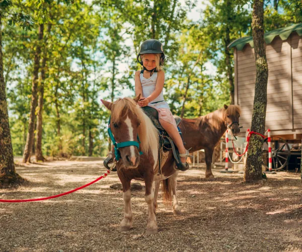 Child riding a pony in a wooded area, wearing a helmet and smiling.