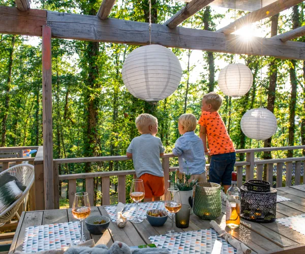 Three children looking outside on a sunlit deck with lanterns and a dining table.