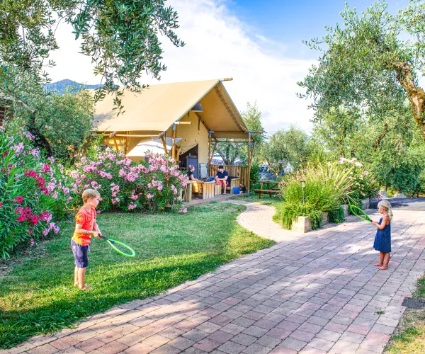 Children playing with rackets near a cottage, surrounded by lush greenery and flowers.