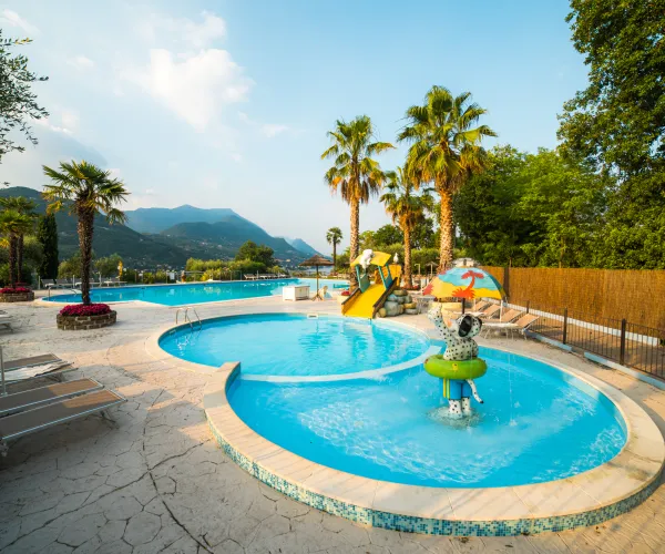 Sunny poolside with palm trees, mountains, and a water slide.
