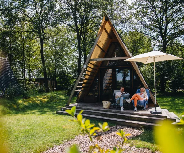 A-frame cabin with two people relaxing on the porch under an umbrella, surrounded by trees.
