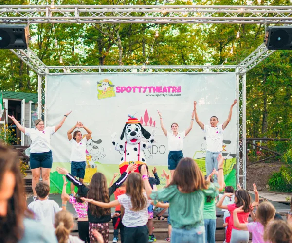 Children dancing on stage outdoors, led by adults in front of a crowd.
