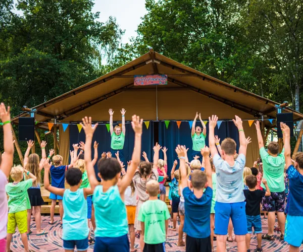 Children raise arms in unison at outdoor event in front of a stage.