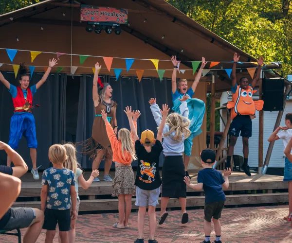 Children and performers on stage, colorful flags overhead, outdoors.