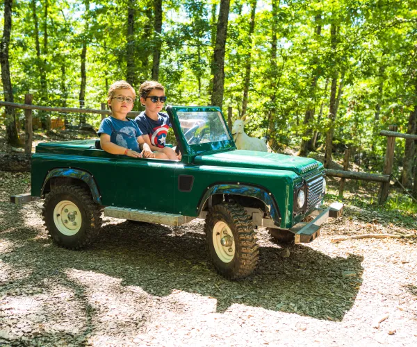 Two kids drive a small green jeep on a forest path, sunlight filtering through trees.