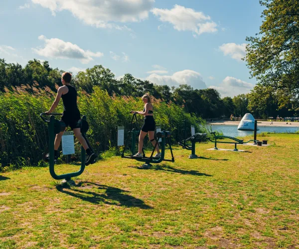 Two people exercise on outdoor stationary bikes by a lake on a sunny day.
