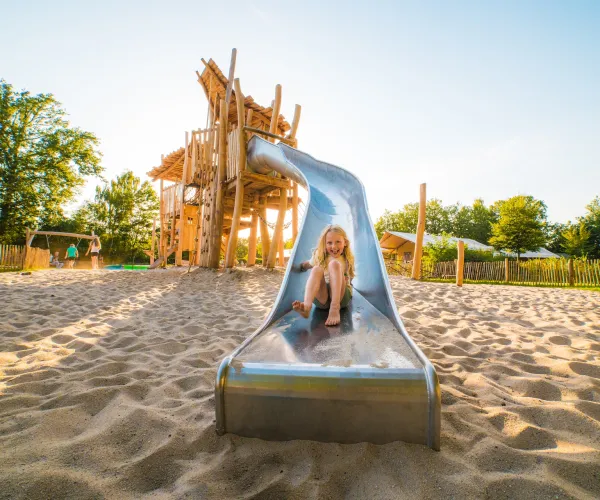 Child sliding down a metal slide in a sunny playground with sand.