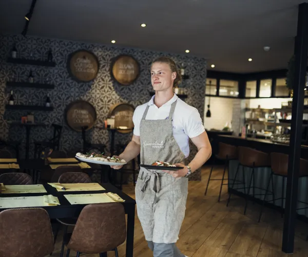Server in apron carrying dishes in a cozy, dimly lit restaurant setting.