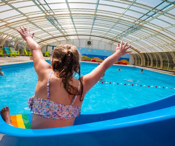 Child on a waterslide in an indoor pool, arms raised in excitement.