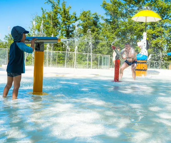 Child in hat playing at a sunny water park with fountains and colorful structures.