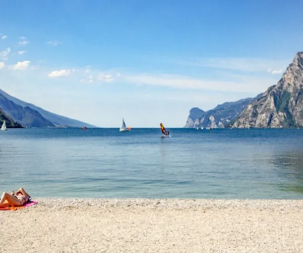 Sandy beach with a person sunbathing by a calm lake, mountains in the background.