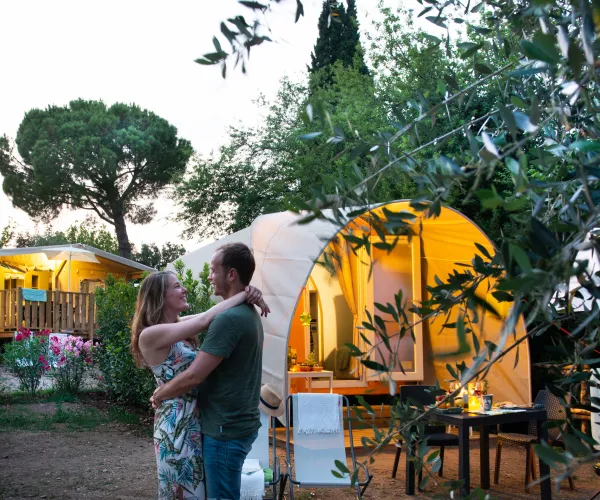 Couple embraces in a garden near a glowing tent at dusk.