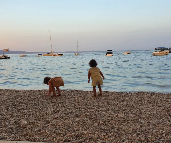 Two children play on a pebbled beach at sunset, boats in the background.