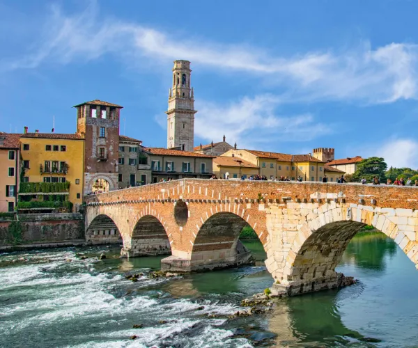 Old stone bridge over a flowing river, with historic buildings and a bell tower.