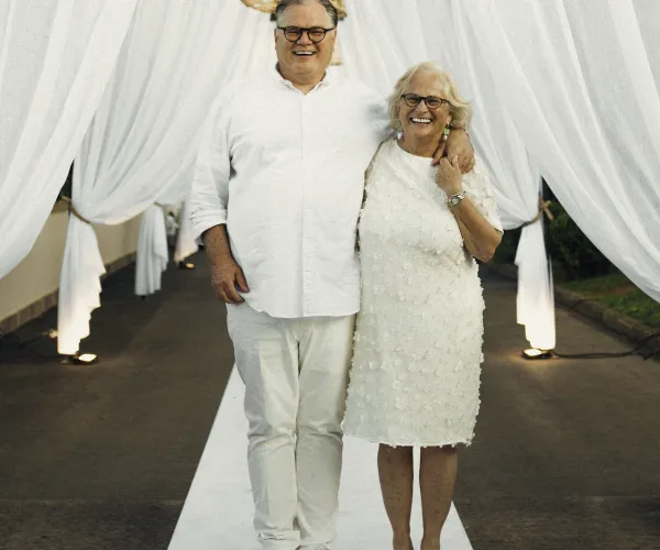 Couple in white outfits standing under draped white fabric outdoors.