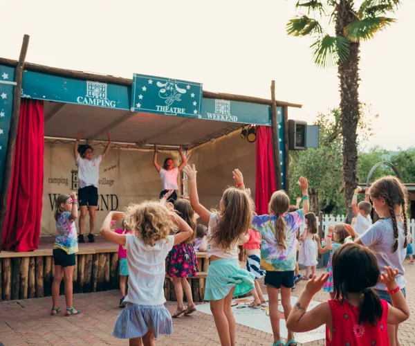 Children dancing in front of an outdoor stage with red curtains.