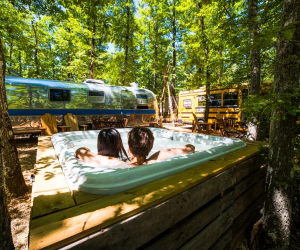 Two people relax in a hot tub surrounded by trees and trailers in a sunny, forested area.