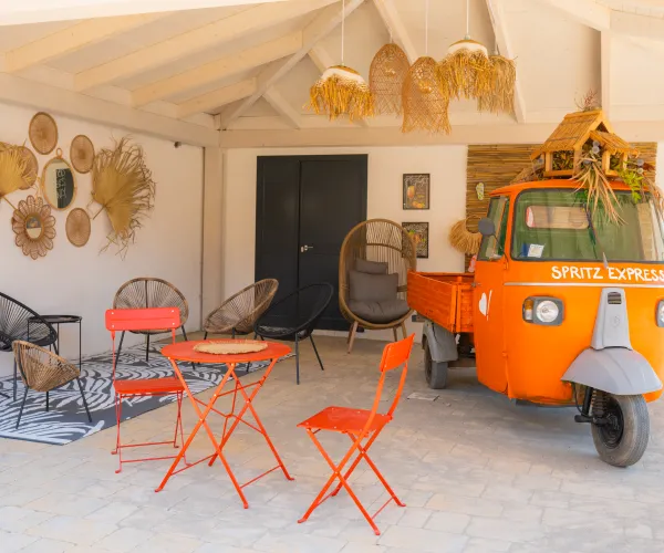 Orange vintage truck in a cozy patio with wicker decor and red metal chairs.